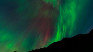 Vibrant aurora borealis lights cascade over a dark, silhouetted mountain range at night