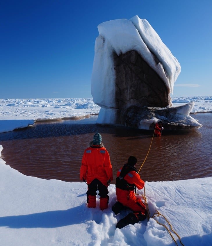 Researchers collect samples using ropes during an expedition aboard the research icebreaker Polarstern (July 2025, expedition PS149). A melt pond has formed at the base of this iceberg locked within Arctic sea ice, tinted brown by sediments contained within the ice