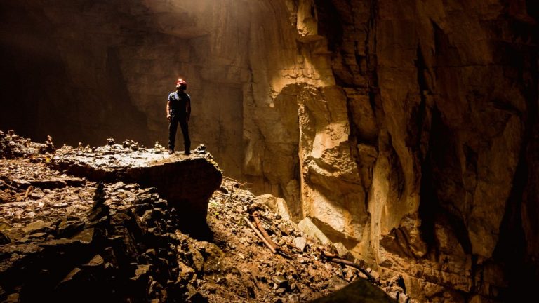 A lone cave explorer stands inside a rocky underground chamber