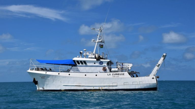A research vessel named La Curieuse sails on calm blue ocean under clear skies