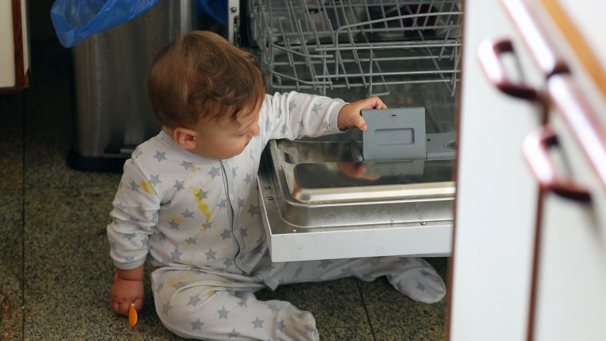 Cute baby touching dishwasher at kitchen. Toddler seated next to machine dishwasher exploring