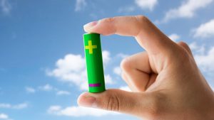 Recycling, energy, power, environment and ecology concept - close up of hand holding green alkaline battery over blue sky and clouds background