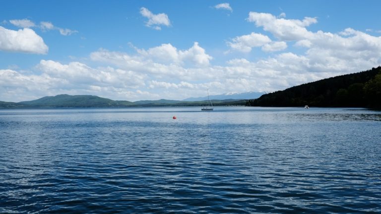 A sailboat crossing a broad lake beneath a blue sky