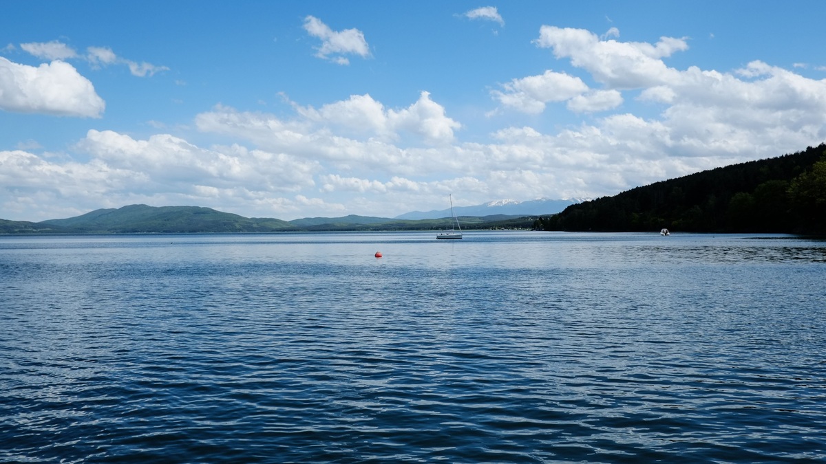 A sailboat crossing a broad lake beneath a blue sky