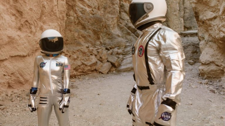Two astronauts in silver space suits exploring a rocky, desert-like environment, resembling Mars