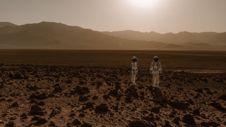 Astronauts in space suits explore a rocky, desert-like terrain resembling Mars at sunset