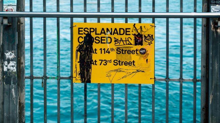 Yellow esplanade closure sign with graffiti on a metal railing by the waterfront in New York City