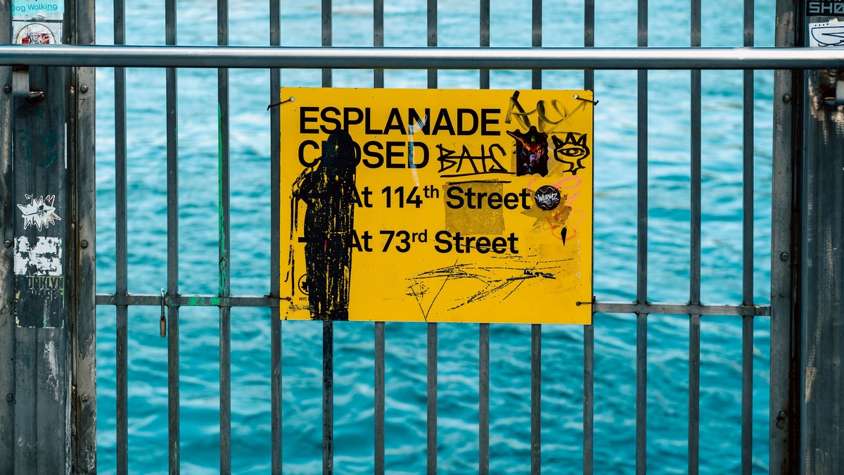 Yellow esplanade closure sign with graffiti on a metal railing by the waterfront in New York City