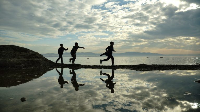 Three children run along the lakeshore in Bursa, Türkiye, silhouetted against the sunrise sky with reflections in the water