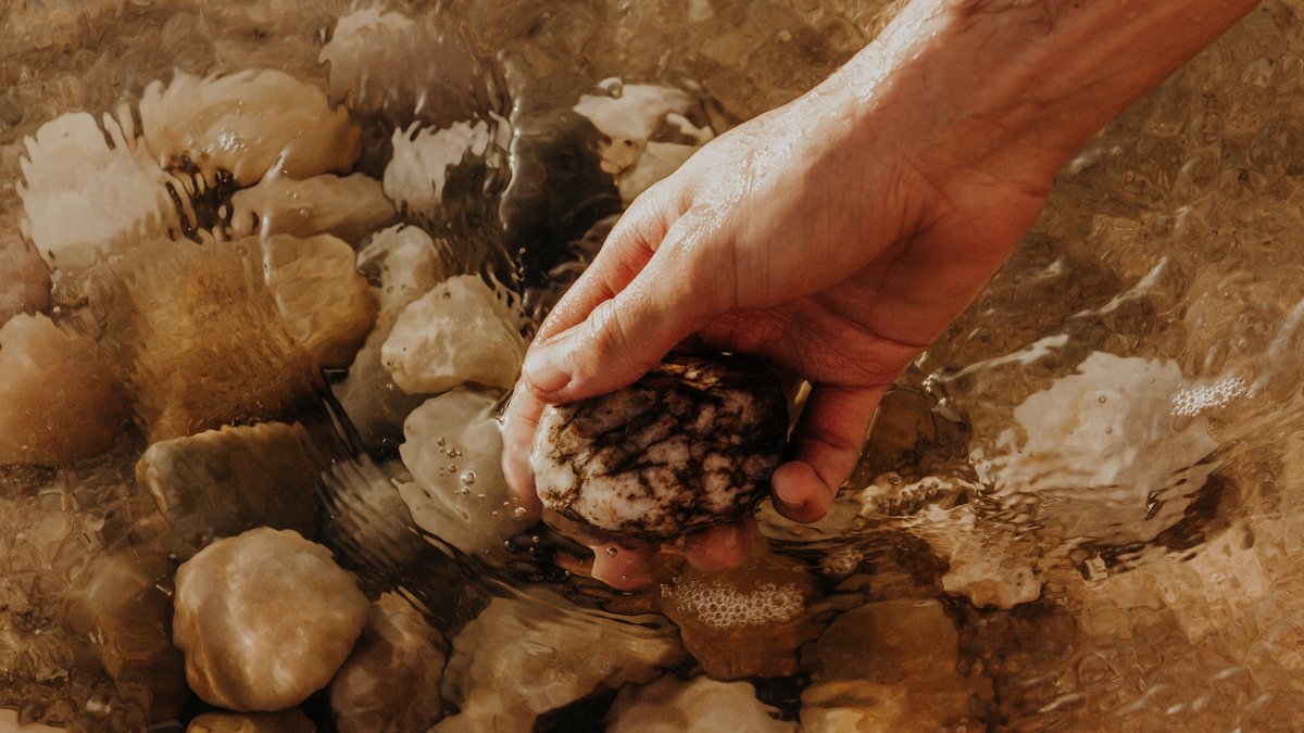 A closeup of a hand holding a stone in shallow water with pebbles