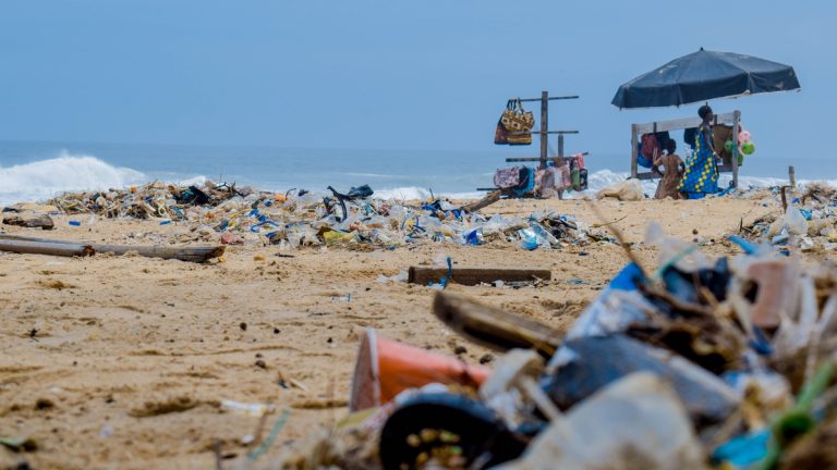 Sandy beach cluttered with trash and ocean debris, with vendors under an umbrella nearby