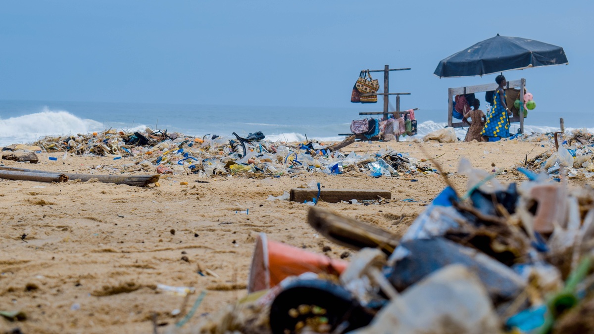 Sandy beach cluttered with trash and ocean debris, with vendors under an umbrella nearby