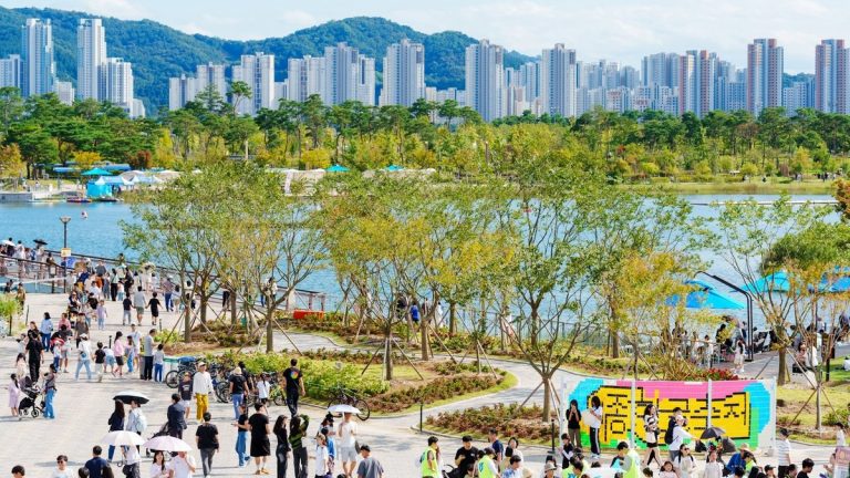 People walking through a busy urban park beside a lake and high-rise skyline
