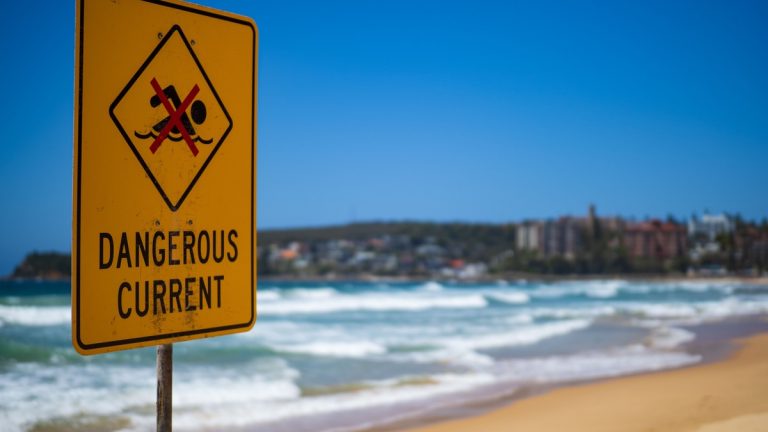 Dangerous current warning sign on Manly Beach in Australia, with blue sky and ocean waves