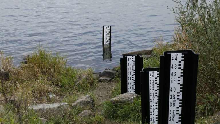 A water level measurement post standing in a lake, surrounded by rocks and vegetation