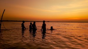 Silhouetted teenagers swimming together in a lake during a vibrant sunset, creating a joyful summer scene