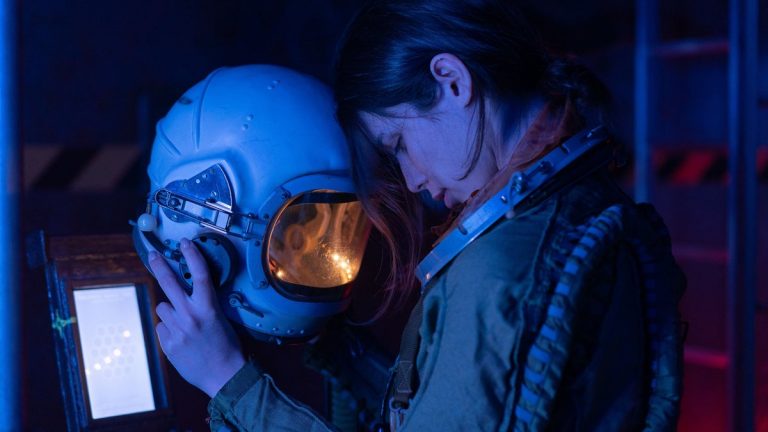 A tired-looking space traveler holding a helmet inside a dim spacecraft
