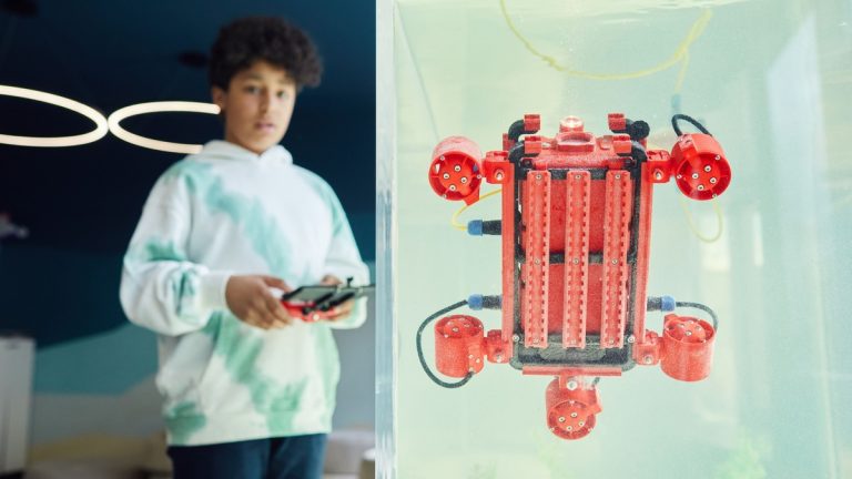 From below of ethnic boy in hoodie controlling robot using panel while standing in light room