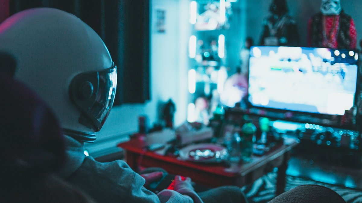 Side view of unrecognizable person in virtual reality helmet sitting on sofa and playing with gamepad in dark room