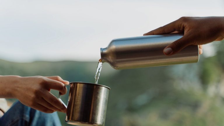 Close-up of hands pouring water from a metal bottle into a cup in an outdoor setting